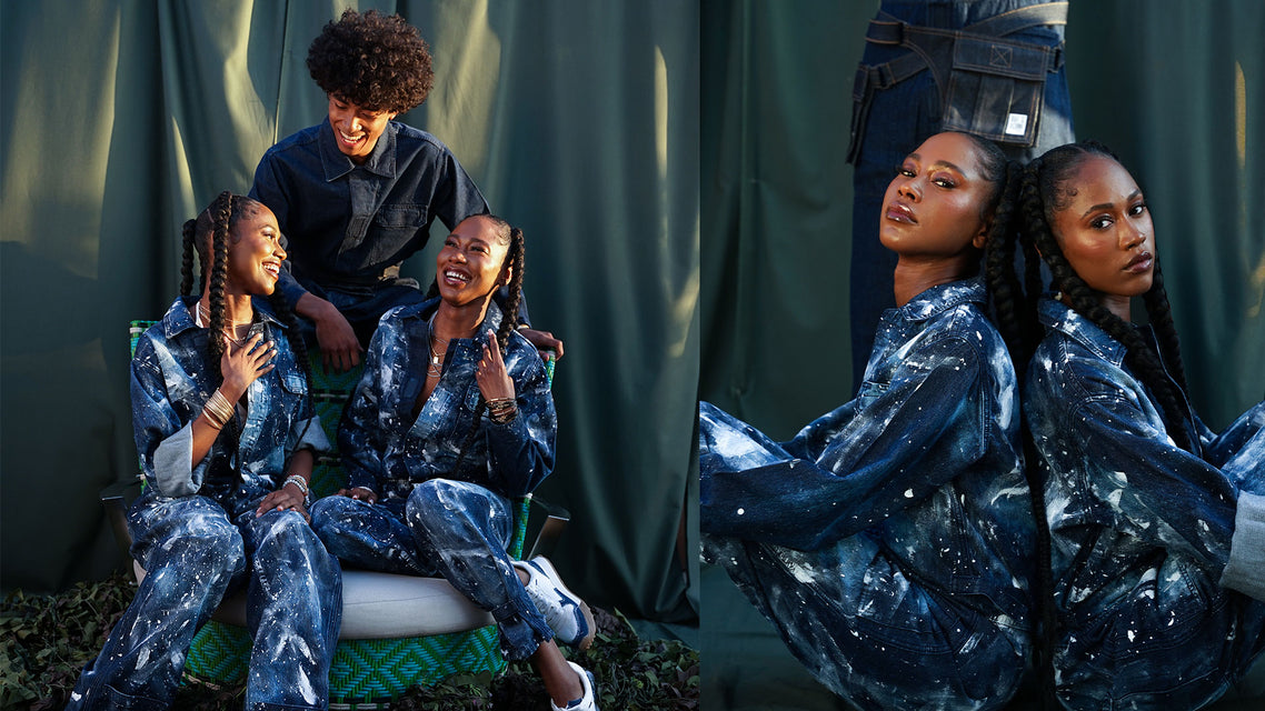 Two young African American Twin sisters sitting side by side laughing and smiling wearing white painted indigo iumpsuits with a young man leaning between them smiling & laughing with a indigo jumpsuit coverall